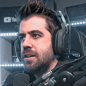 AuronPlay, famoso creador de contenido español, con barba y cabello oscuro, vistiendo una chaqueta negra y una camiseta blanca, sonriendo mientras mira hacia un lado.