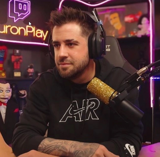 AuronPlay, famoso creador de contenido español, sonriendo mientras transmite en vivo desde su setup gamer, con auriculares, micrófono y luces LED de fondo.